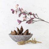 A boat-shaped stone bowl with flower detailing to the surface, shown holding dried sorocca flowers, displayed alongside gold canella and faux pink eucalyptus