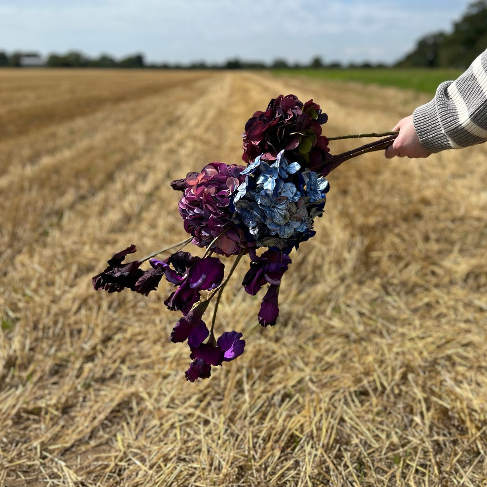 A group of deep red and metallic blue Silk-ka artificial flowers, held up against a stubble field.