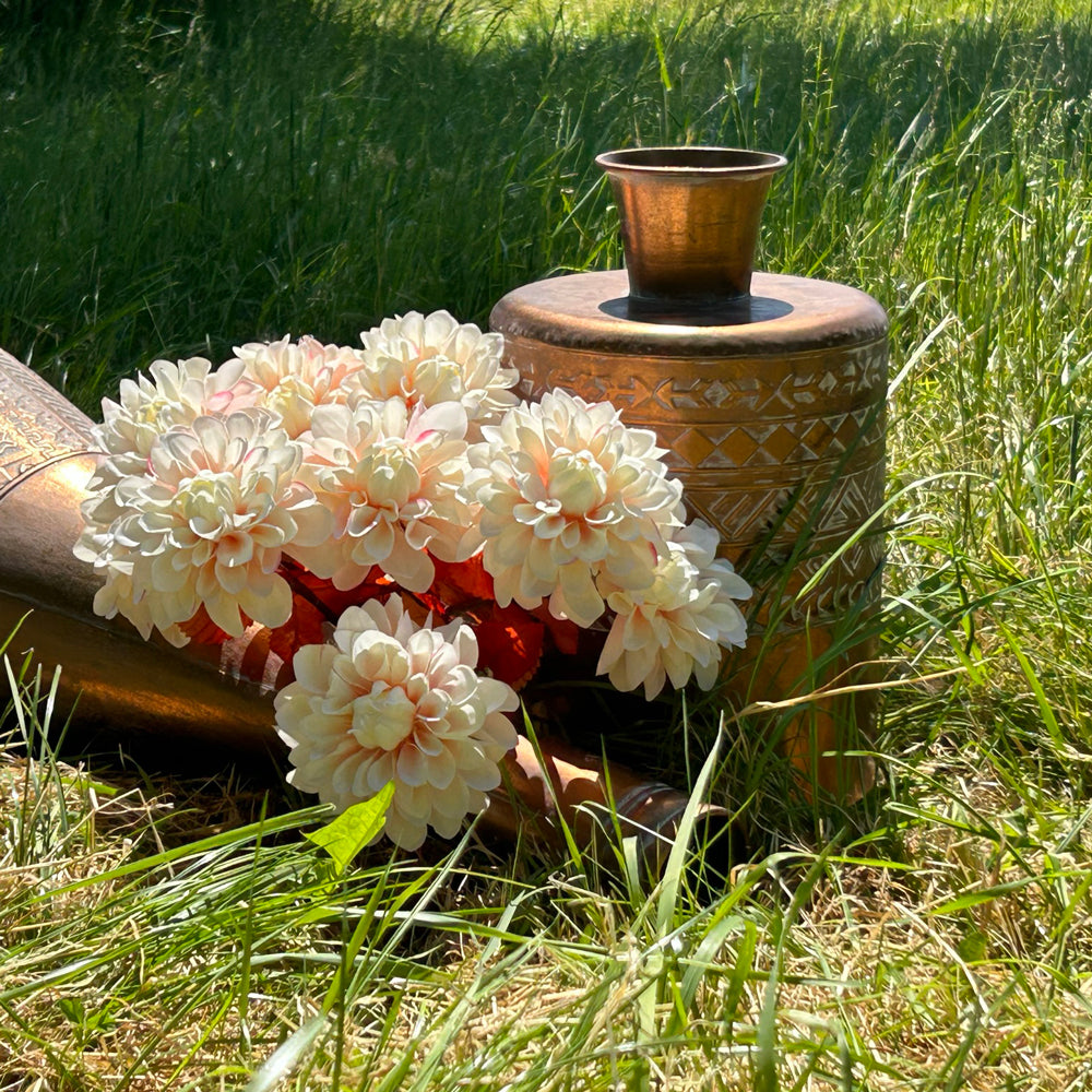 A group of faux, light pink dahlias displayed with copper coloured vases, within a meadow on bright sunny day