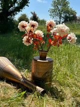 A group of faux, light pink dahlias displayed with copper coloured vases, within a meadow on bright sunny day