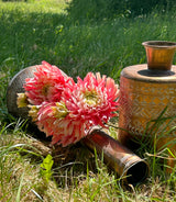 A group of fluffy pink chrysanthemums with orange leaves, displayed in a copper vessel, against the backdrop of a meadown in bright, summer sunshine.