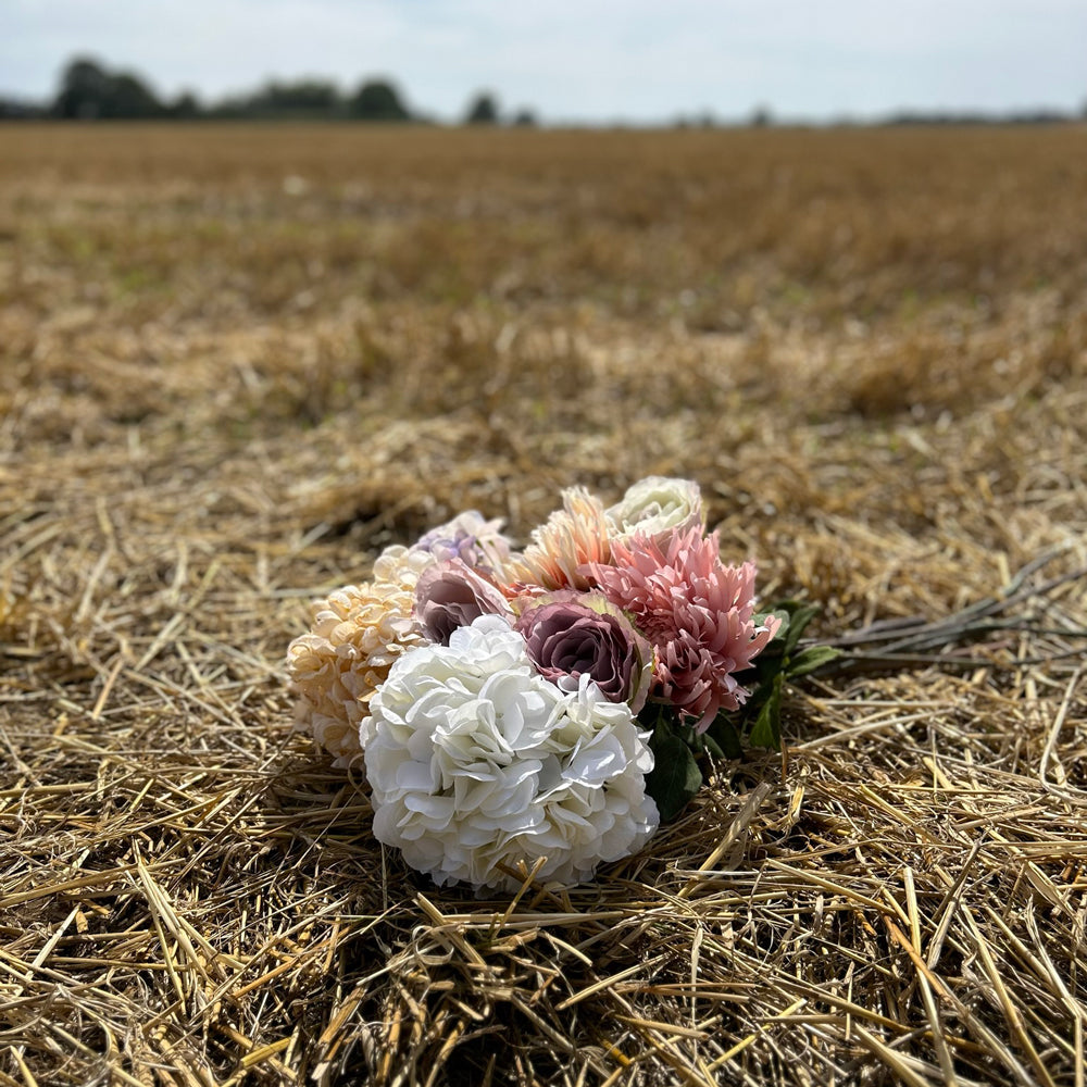 A collection of pastel coloured Silk-ka faux flowers, held against a cloudy sky and stubble field.
