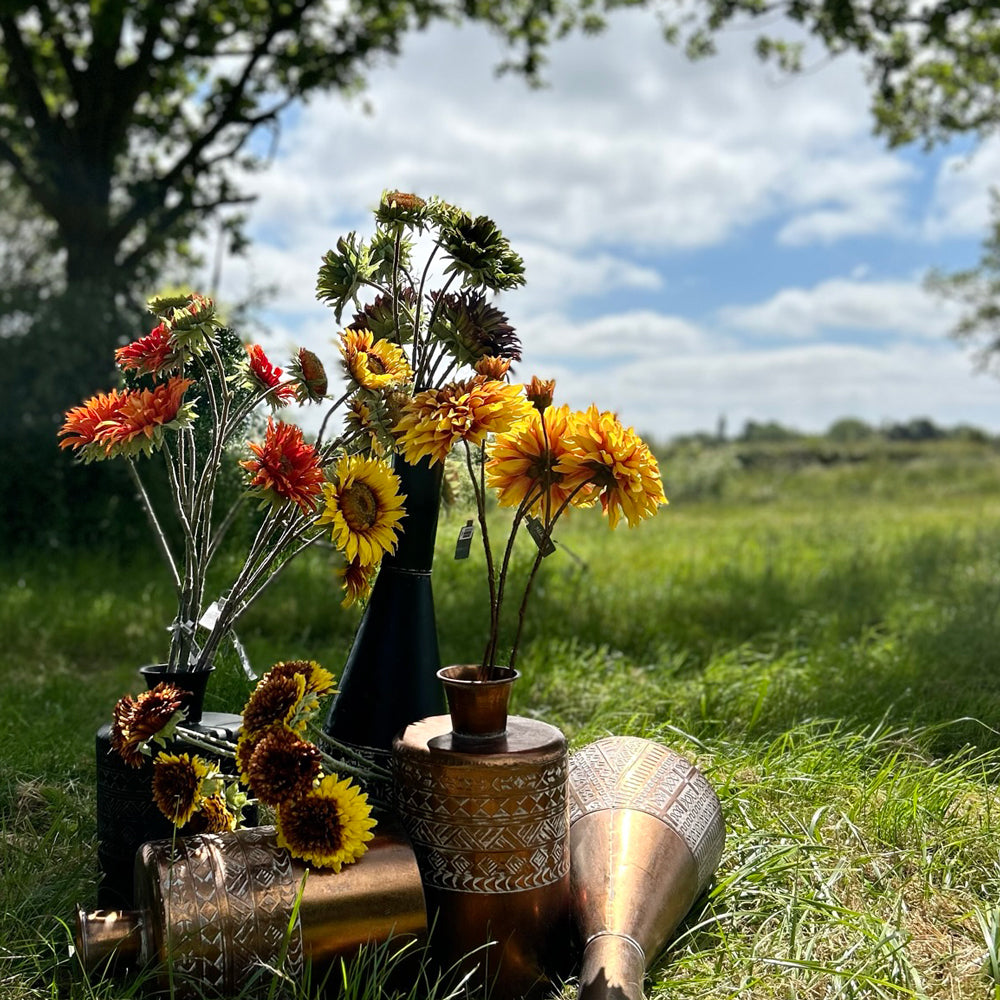 A group of different coloured, sized, and shaped faux sunflowers, displayed in a meadow against a bright, sunny sky
