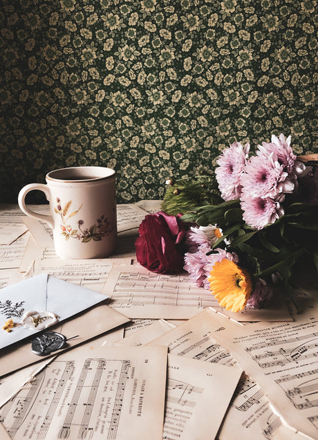 A table covered in music sheets, flowers, and with a dark green floral wallpaper as a backdrop.
