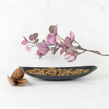 A dark grey, canoe-shaped , flat bowl, containing gold-painted Casuarina Pods. Also shown are pink, faux eucalyptus and a single, dried sorocca flower/