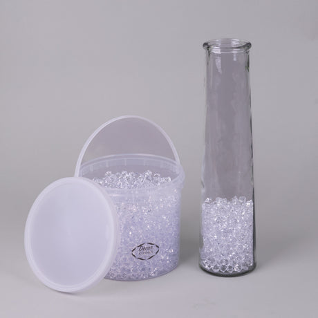 An open bucket of small, clear acrylic stones, in a plastic tub, next to a tall clear vase with some of the stones inside as an example.