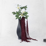 Burgundy amaranthus in a black vase with faux white lilies and green foliage.
