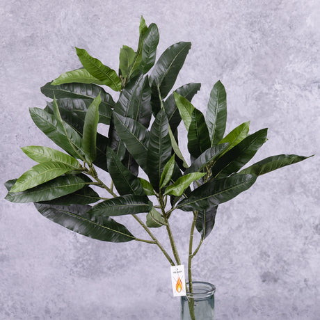 A faux mango bush stem showing lots of deep green leaves, displayed in a clear glass vase showing the fire retardant label