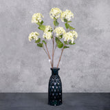 Three faux viburnum, or snowball flowers, in a cream colour with green tinges, displayed in a blue glass vase