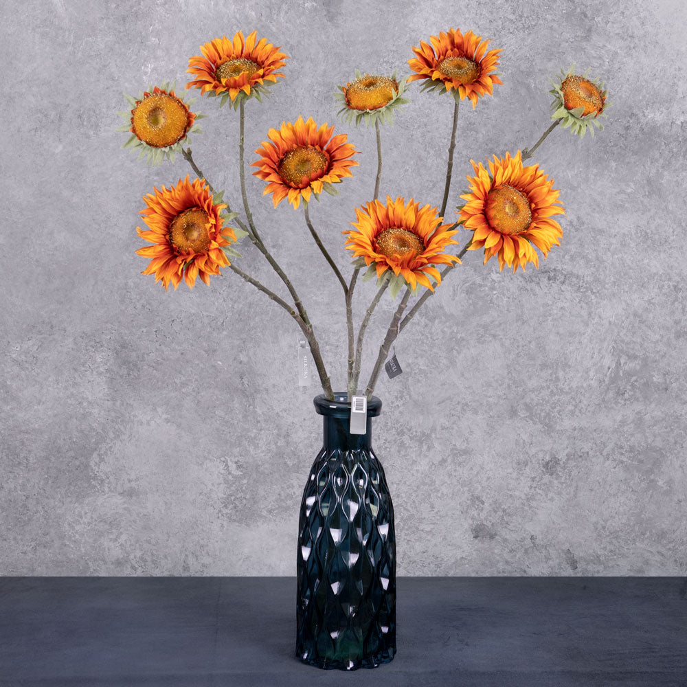 A group of three faux sunflower sprays in a rich orange colour, displayed in a blue glass vase