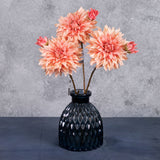 A group of three faux dahlia sprays with rich, salmon-coloured flowers and emerging buds, displayed in a blue glass vase