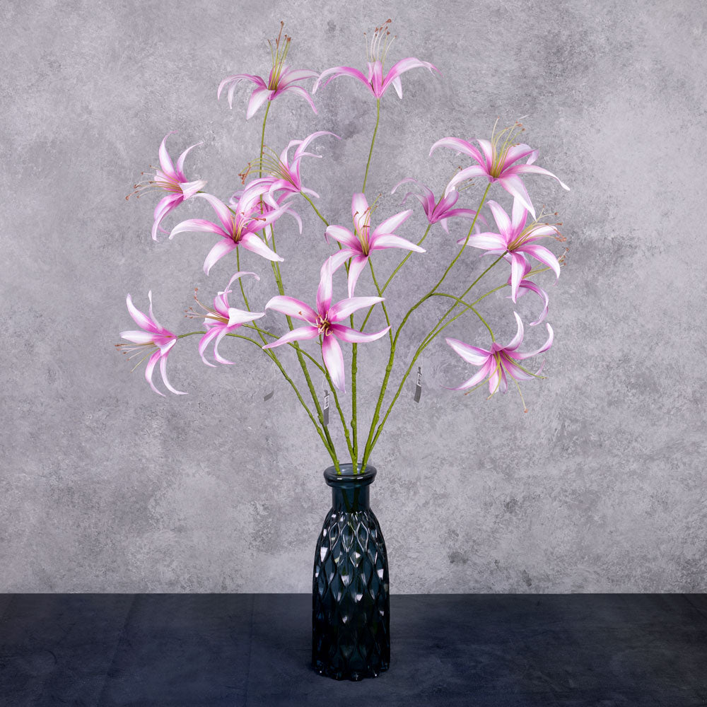 Three tall. faux lily sprays with light pink flowers that have darker pink edges to the petals, shown in a dark blue glass vase