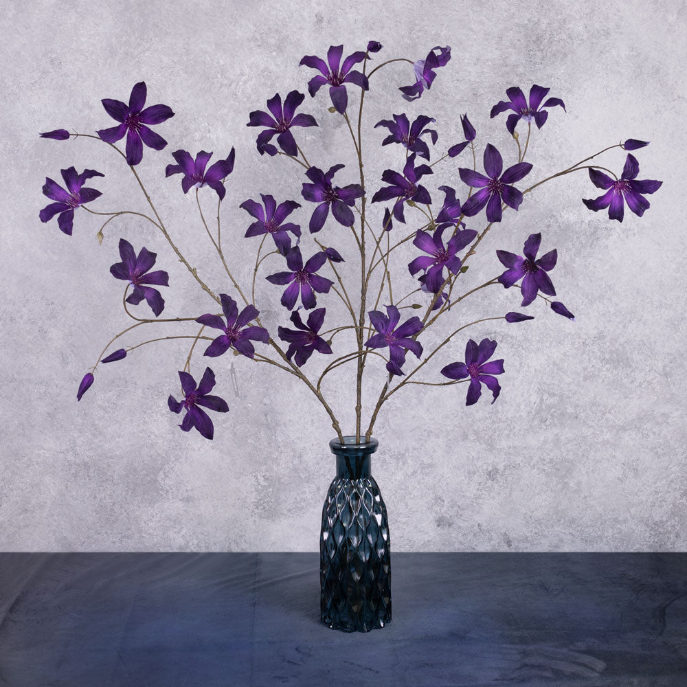 Three faux clematis sprays with deep purple coloured flowers, shown in a blue glass vase