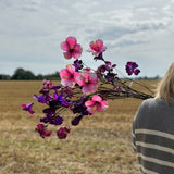 A group of pink and purple faux flowers by Silk-ka, held up against a slightly cloudy sky and a large stubble field.