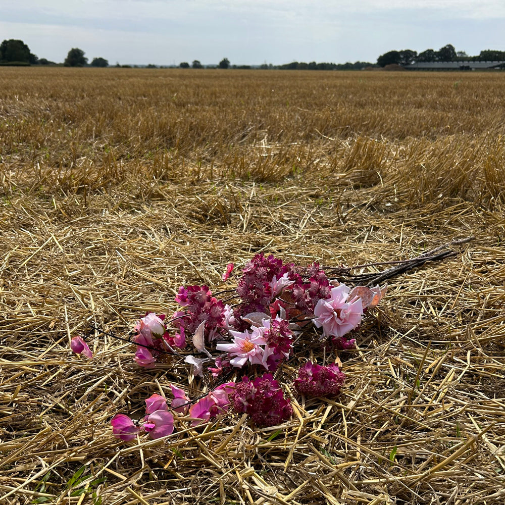 A bunch of different, pink, artificial flowers laid on a stubble field with an overcast sky in the background.