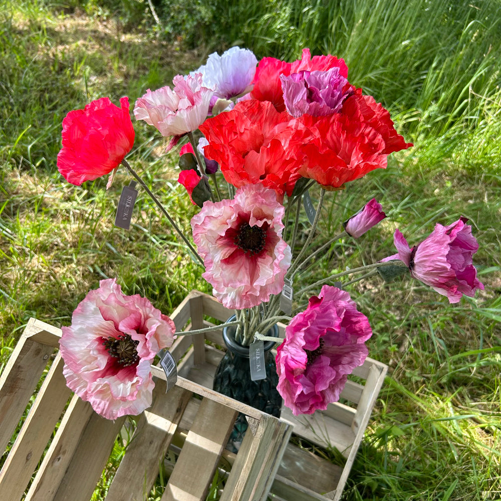 A group of faux poppies in reds, pinks, lilac and purple, against a meadow, trees and bright blue sky