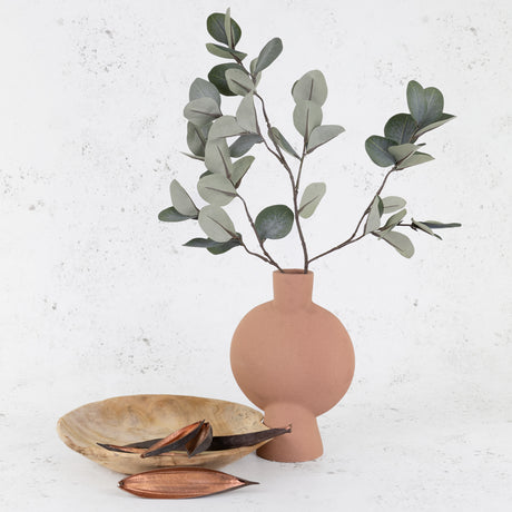 A hand made porcelain vase containing a faux eucalyptus stem, next to a hand-carved teak bowl holding decorative coco boats