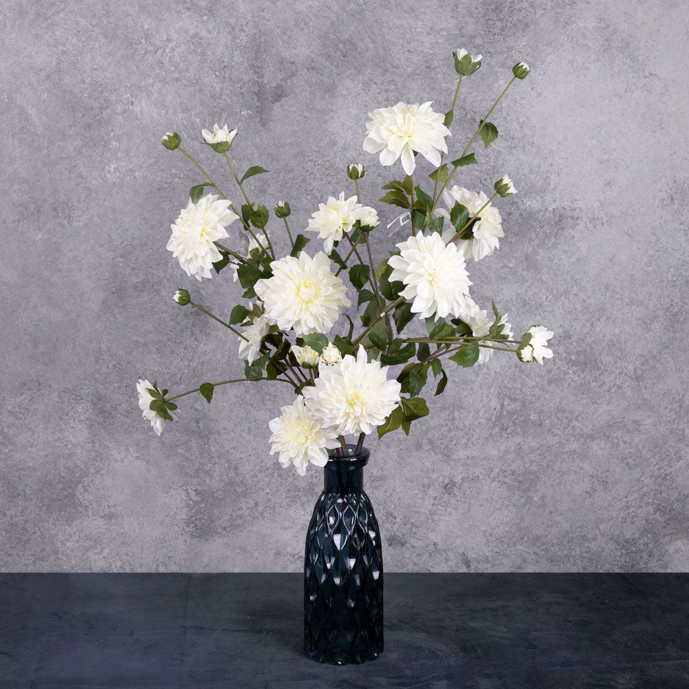 Three white, faux dahlia sprays, with open flowers and buds, in a blue, glass vase