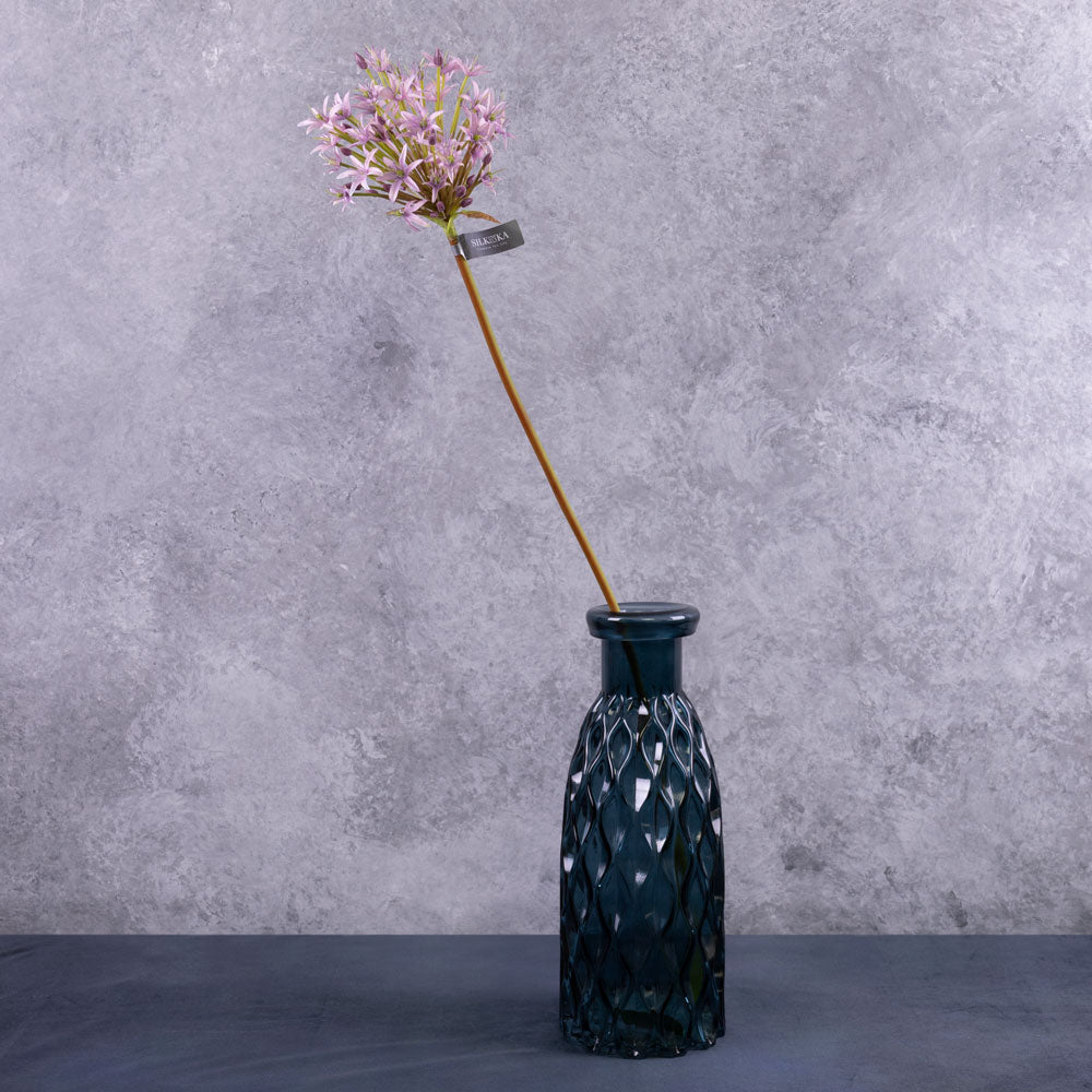 A single faux allium stems with a lilac coloured flower plume, displayed in a blue glass vase