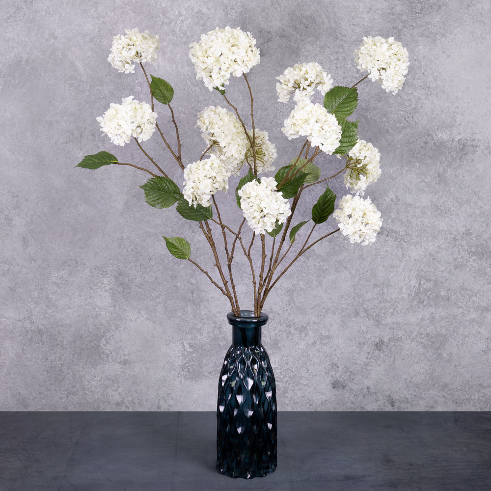 3 faux hydrangea sprays with four, pom-pom-like cream flowers, and five leaves per sprray, displayed in a blue glass vase.