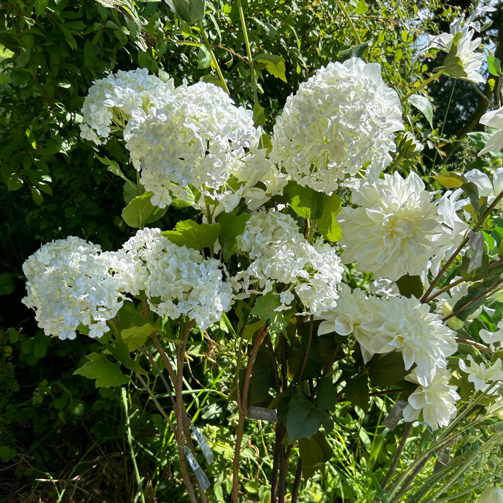 3 faux hydrangea sprays with four, pom-pom-like cream flowers, and five leaves per sprray, displayed in a sunny meadow location against a tree and long grass