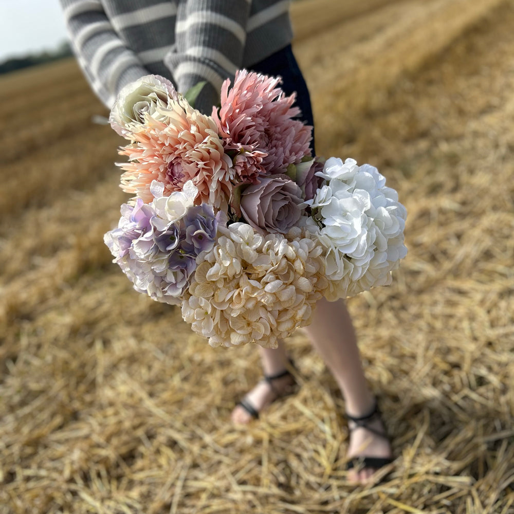 A collection of pastel coloured Silk-ka faux flowers, held against a cloudy sky and stubble field.