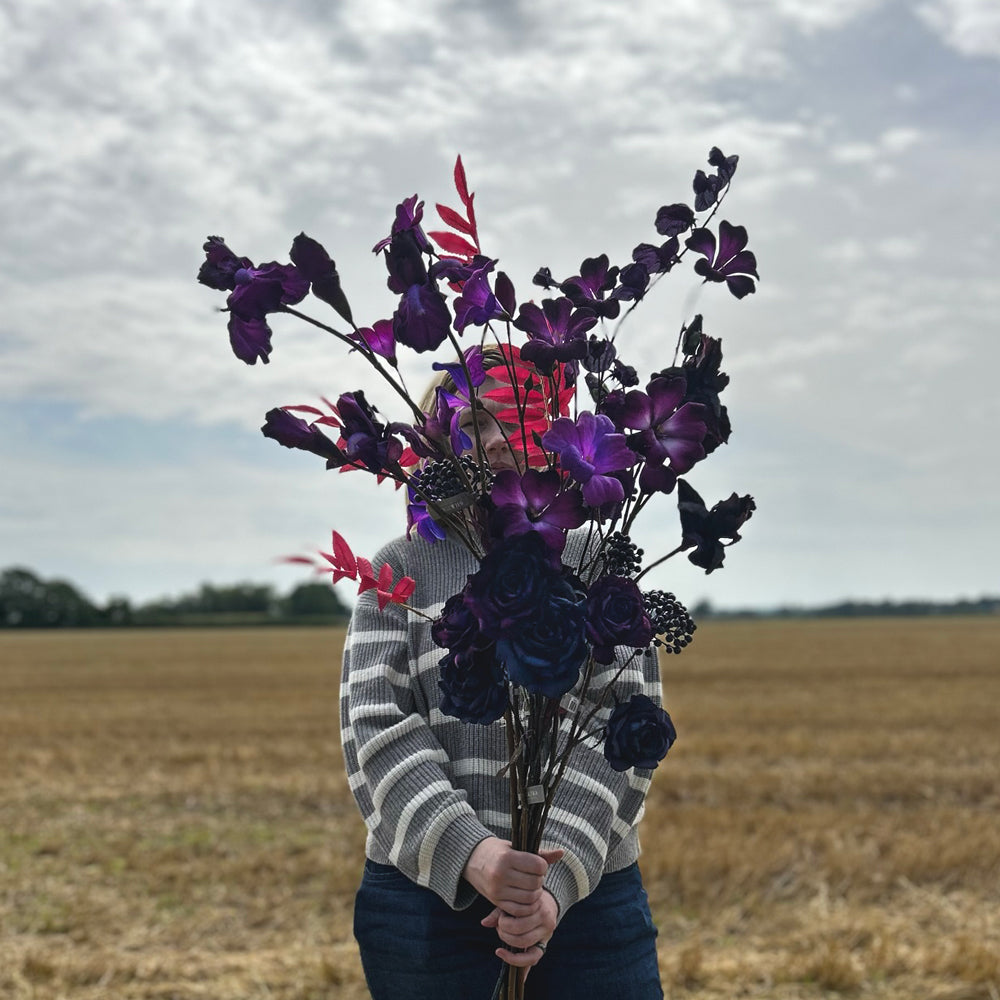 A collection of long-stemmed Silk-ka faux flowers and berries in a range of purples and deep pinks, held up in front of a cloudy sky and stubble field.